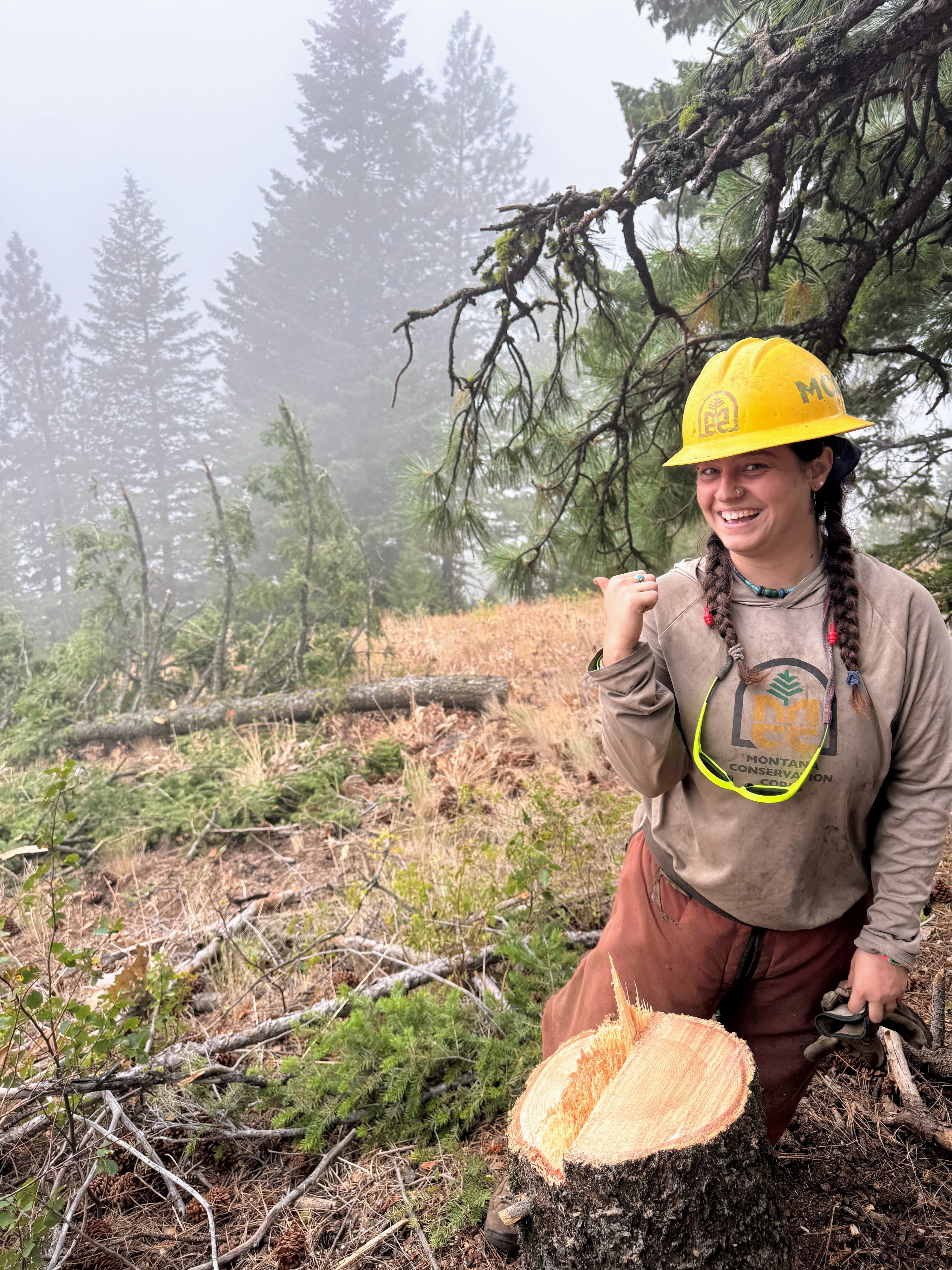 A crew member stands on a hillside and smiles pointing to a tree they cut down.