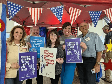 Five individuals are standing together in front of a red canopy decorated with American-themed banners and bunting featuring stars and stripes. They are smiling and holding signs promoting voting rights with quotes from figures like Susan B Anthony and La