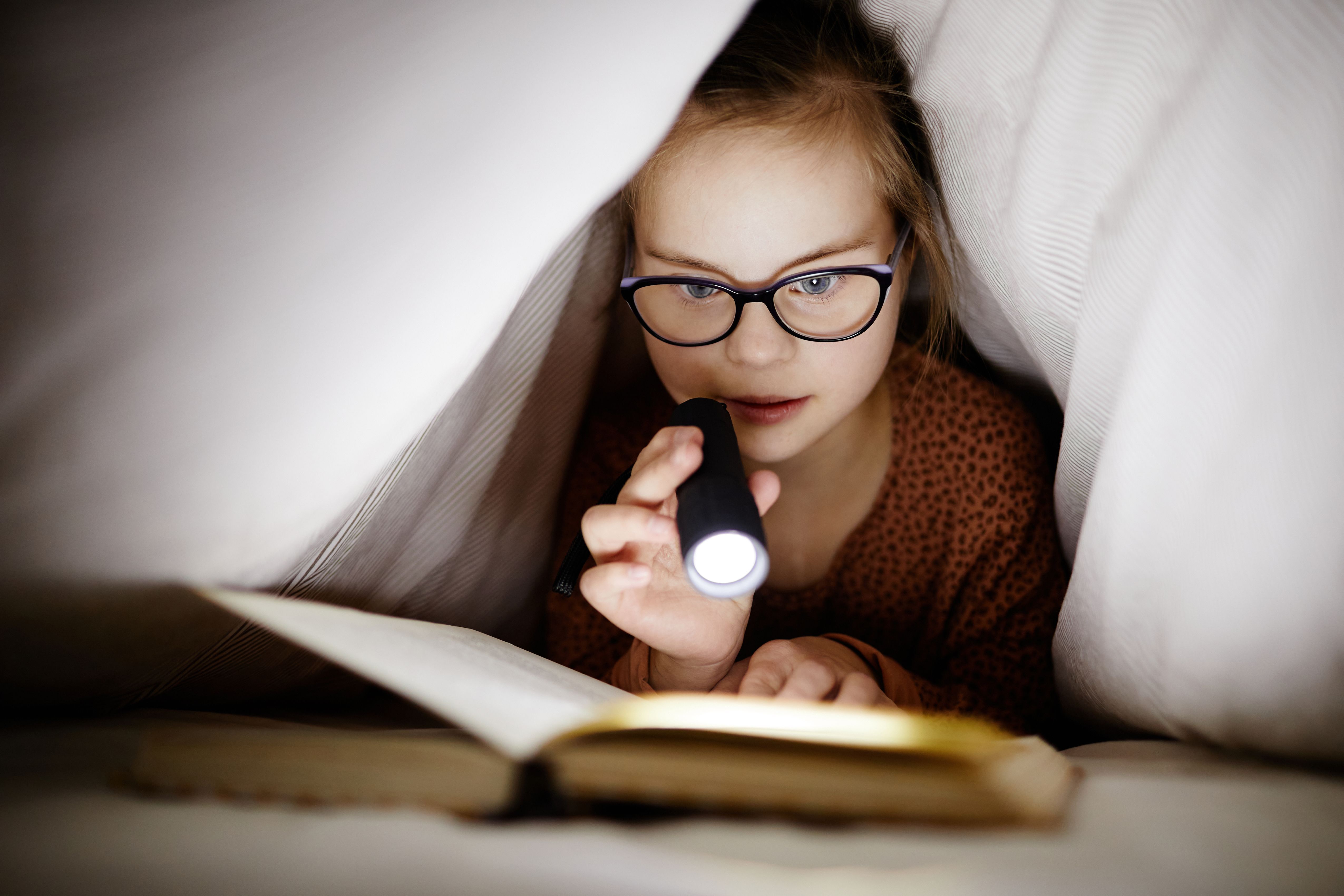 Girl with down syndrome reading a book under covers with a flashlight.
