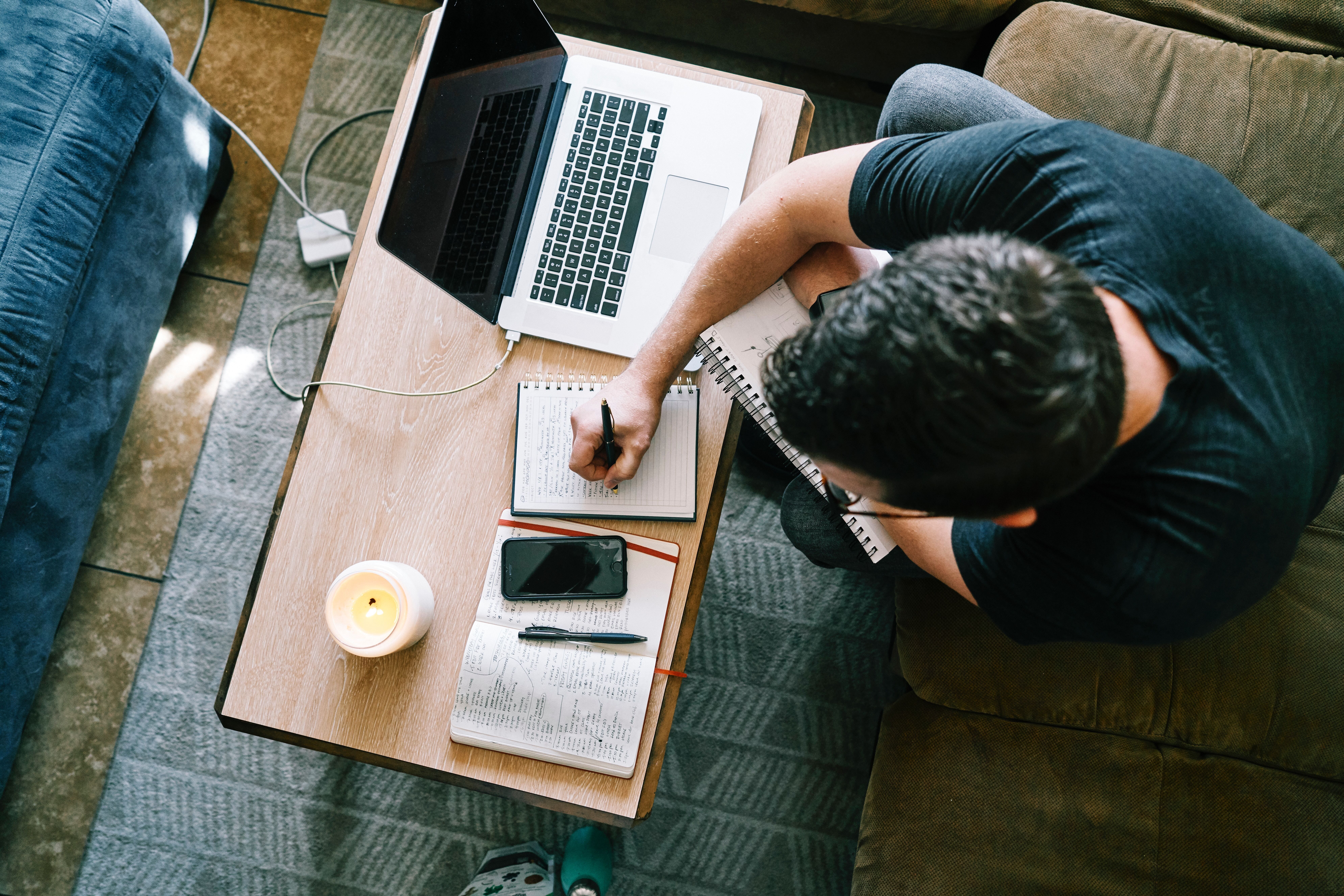 Guy with gray t-shirt writing at table. The table has tablet, notebook, and candle. There is also a charging cord going to the laptop.