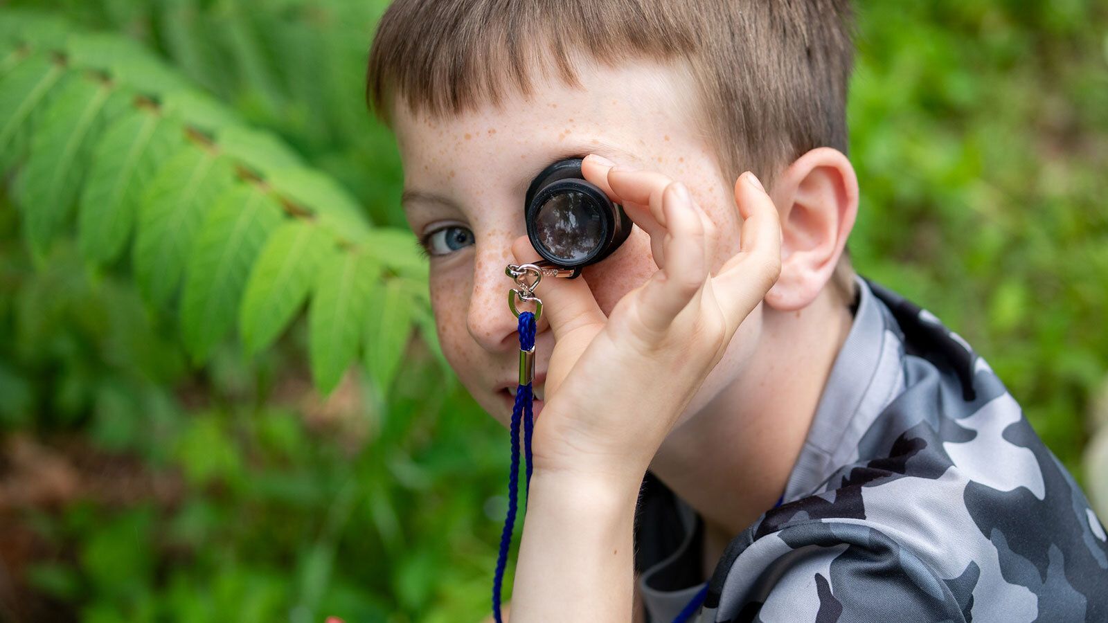 Child looking through a magnifying glass outdoors near green ferns.