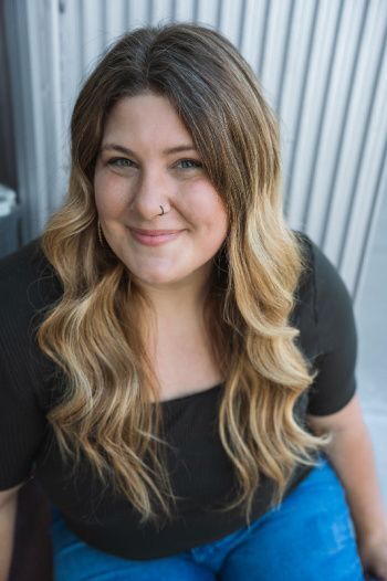 Smiling young woman with long wavy blonde hair dark at the roots and a nose ring, wearing a black scoop-neck top and blue jeans. She sits against a vertical corrugated metal backdrop, looking up at the camera with a friendly, confident expression.