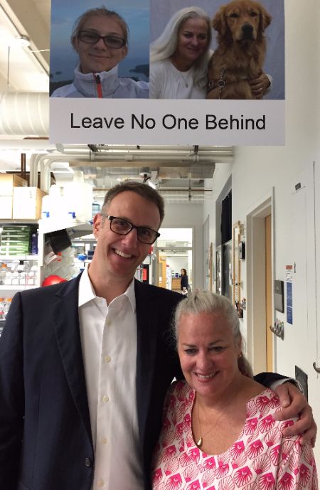 Photo of Moira Shea and Mark Dunning at the Usher Syndrome Research Lab in Iowa, in front of a sign with pictures of Bella Dunning and Moira and her dog that reads "Leave No One Behind"