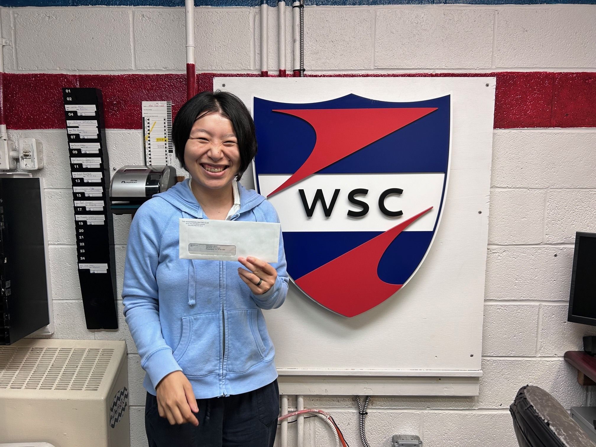 Miranda holding her first paycheck in front of the Wissahickon Skating Club logo.