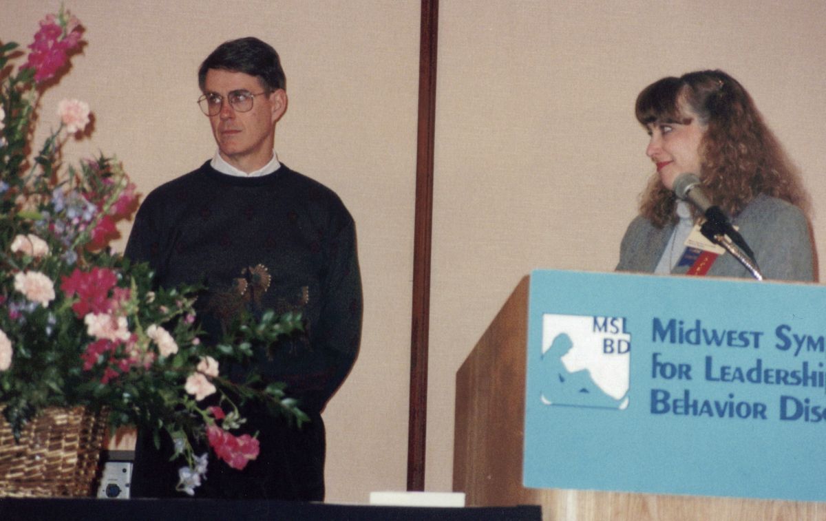 Old photo of man in blue sweater and glasses standing beside a woman who is standing in front of a podium looking at the man beside her. 