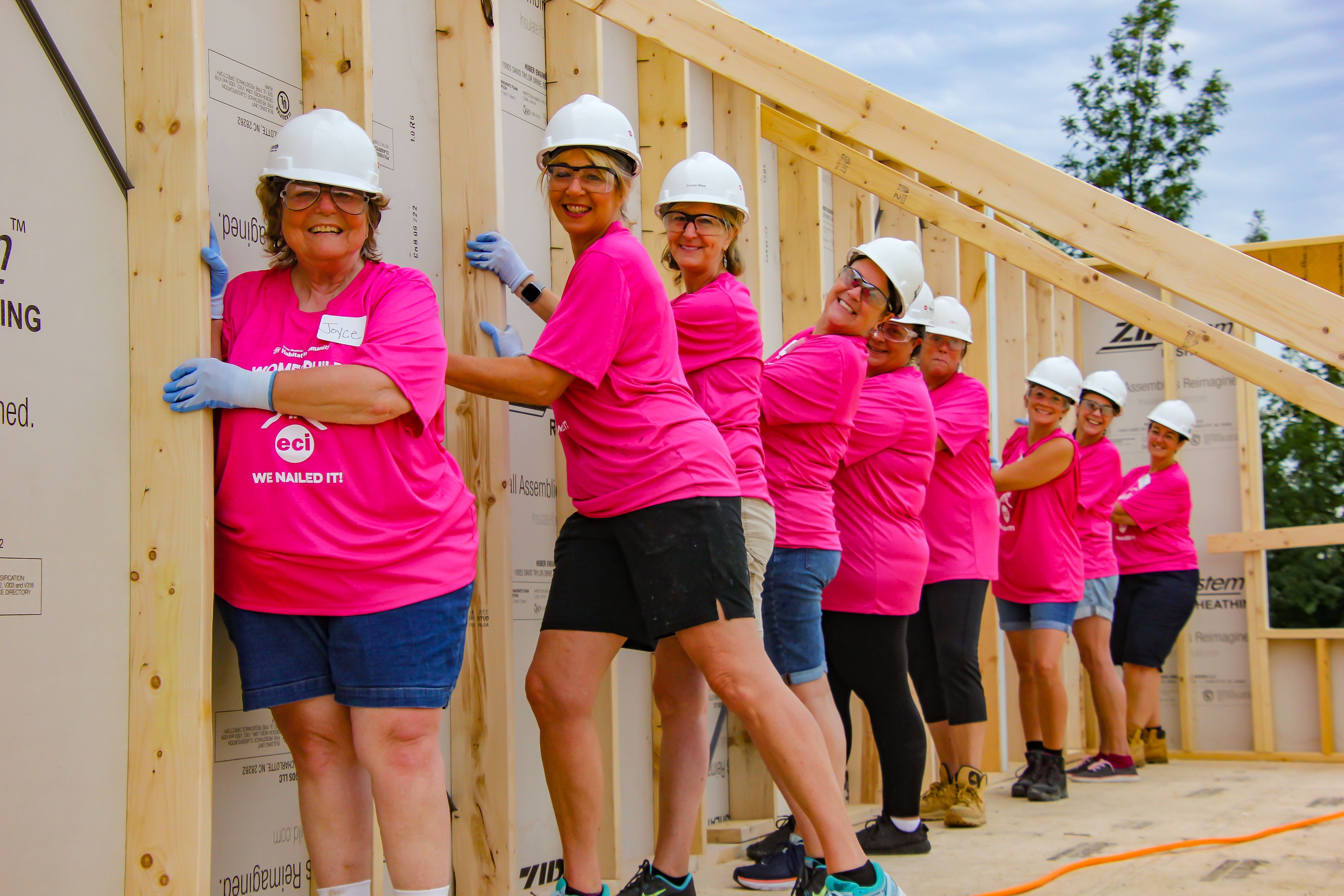 Women in pink shirts and hard hats help raise a wall.