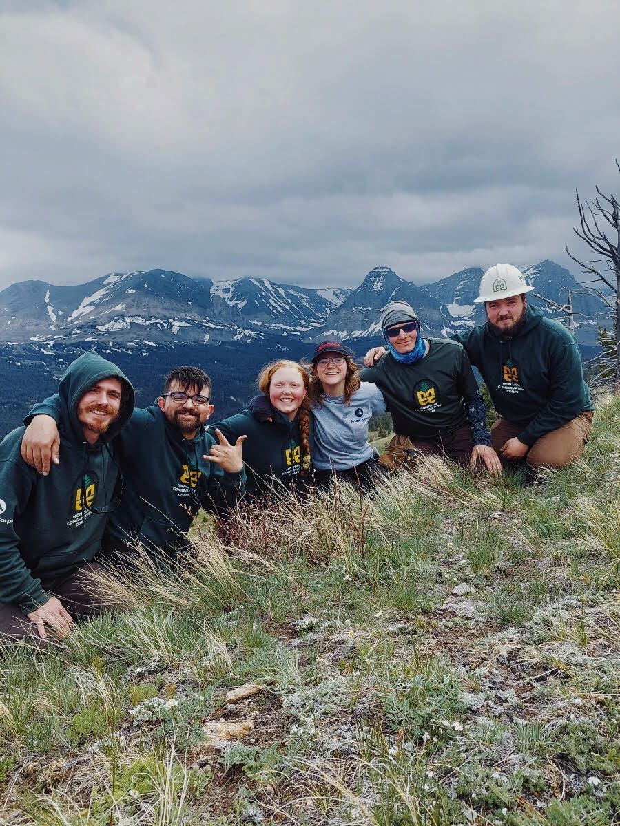 A crew is crouched, smiling towards the camera, in front of a beautiful mountain vista.