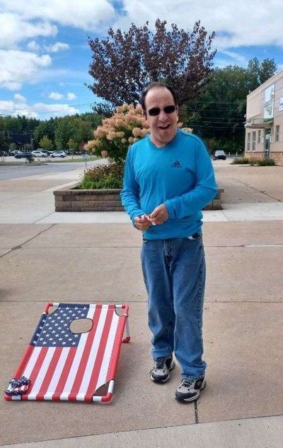 A man faces the camera with a bean bag in his hand.  He stands next to a cornhole board.