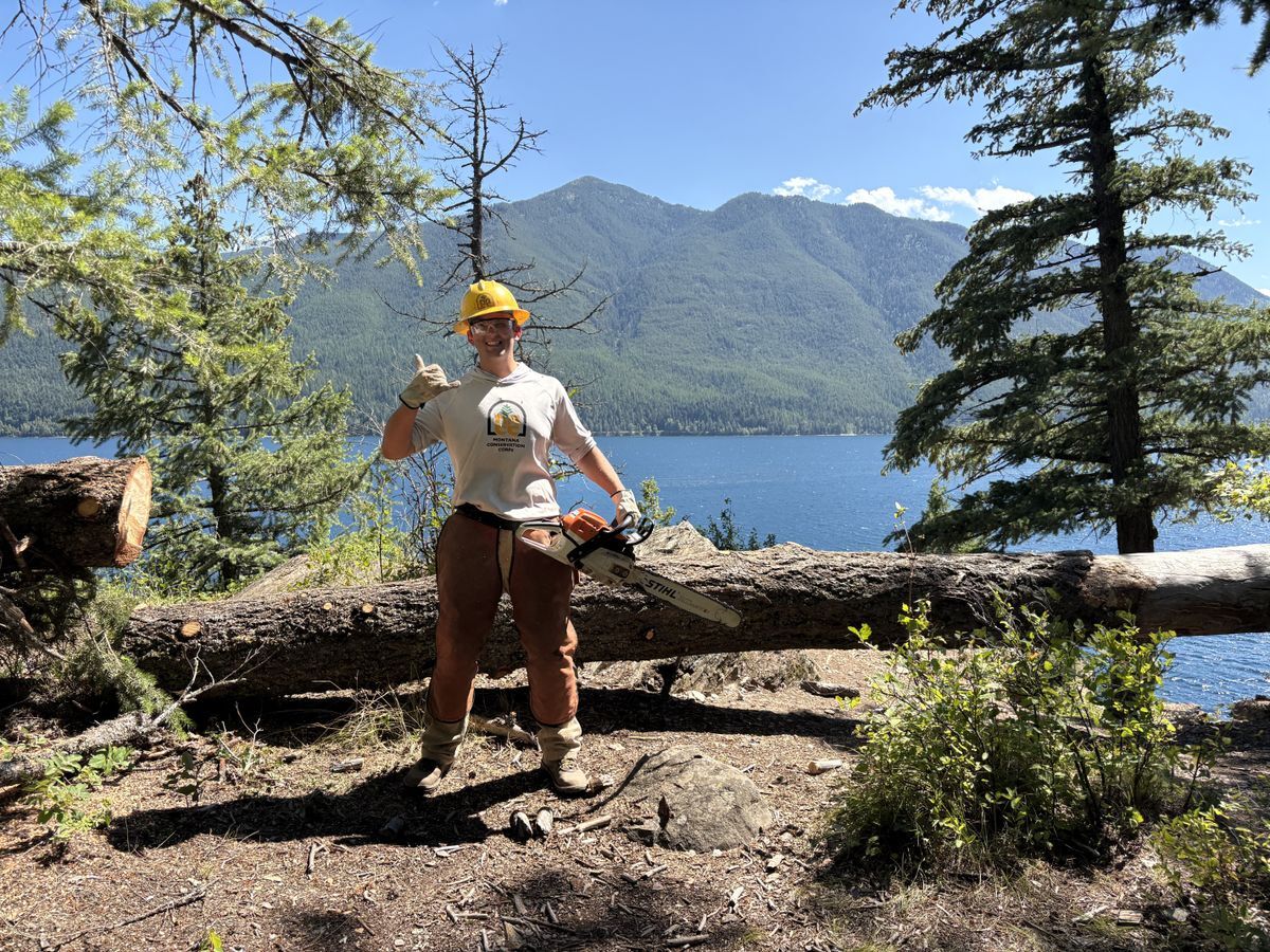 [Image Description: An MCC crew member holds a chainsaw in front of a log, smiling towards the camera and giving a thumbs up.]