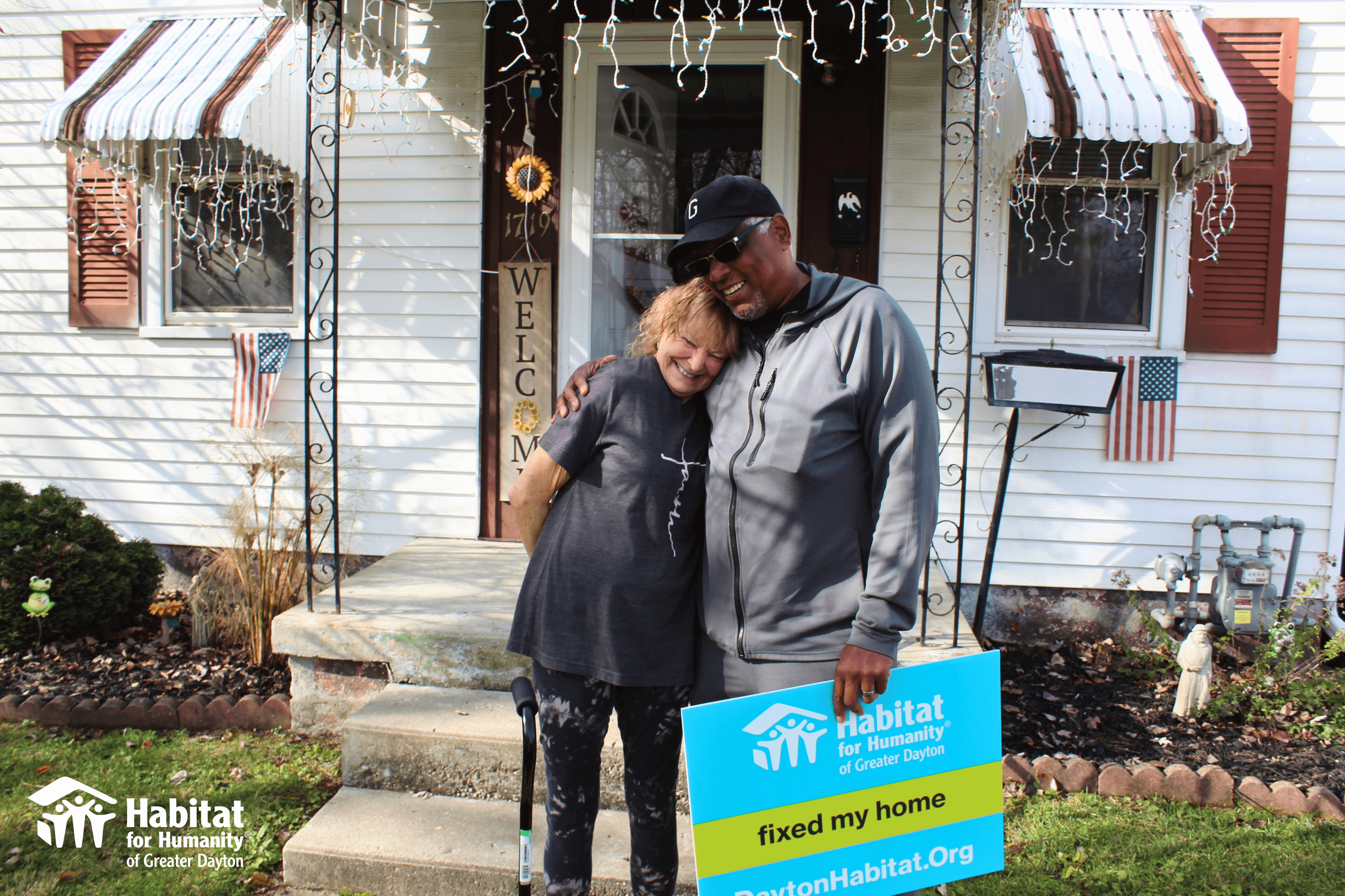 A women hugs a man who just helped fix her home. They are standing in front of her home holding a sign saying "Habitat fixed my home."
