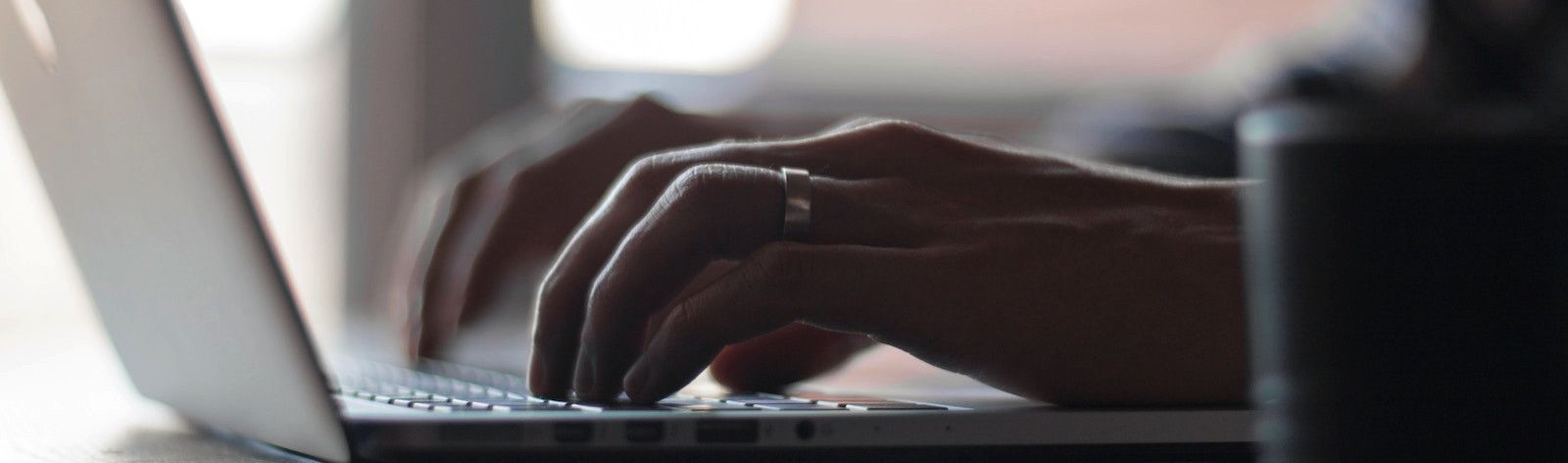 Sideview photo of a man's hands poised to type on the keyboard of a laptop. The laptop is silver and the man wears a wedding band on his left ring finger.