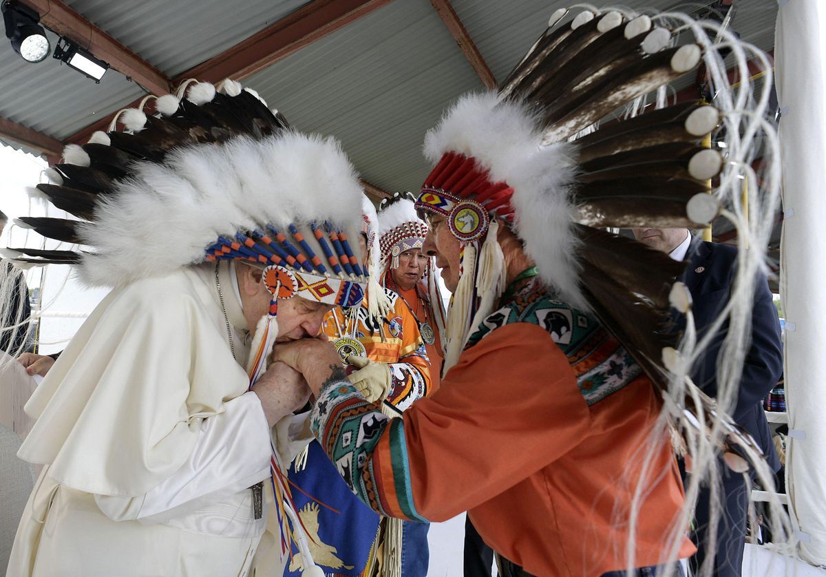 Pope Francis bows and kisses the hand of an Indigenous man, while both wear headdresses.