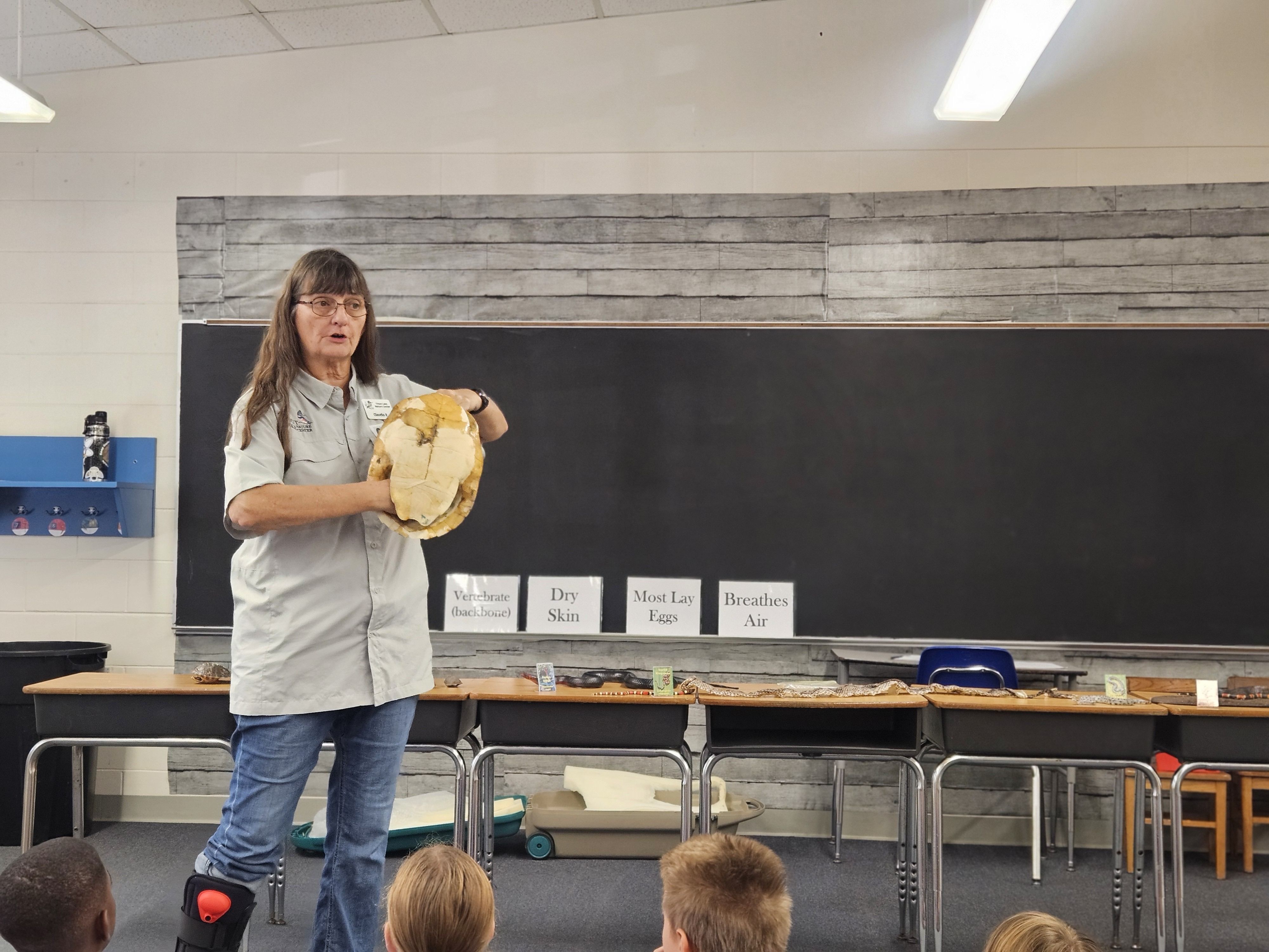 TLNC staff member holds a turtle shell at the front of a classroom. One the white board behind her are listed the characteristics of a reptile.