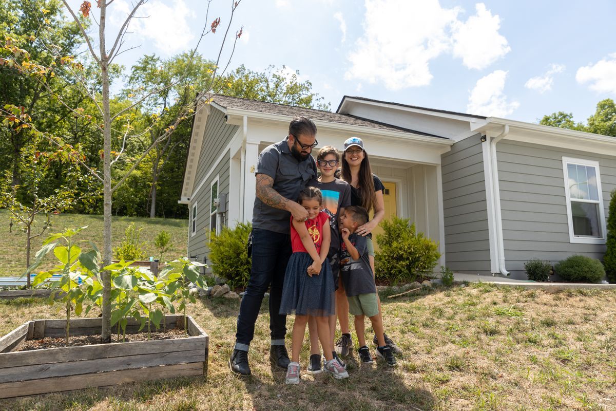 A family of five stands together in front of their Habitat for Humanity home on a sunny day, smiling and hugging in a green yard with a raised garden bed nearby.
