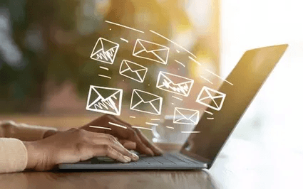 A person is typing on a laptop with white illustrated envelopes floating above the keyboard symbolizing email activity. The background is softly blurred with warm lighting creating a professional and dynamic atmosphere.