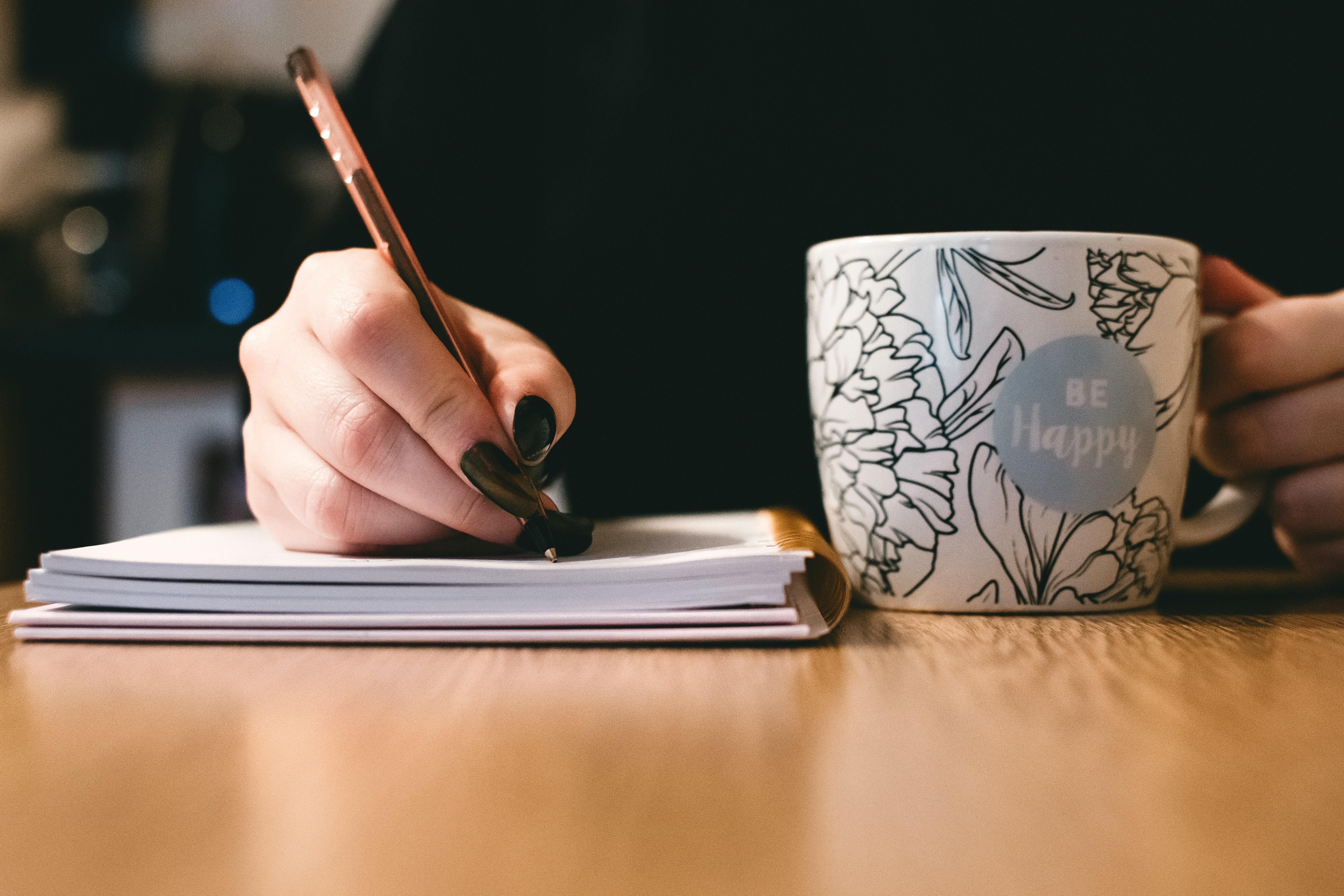 A closeup of a woman's hand writing in a notebook next to a coffee mug.