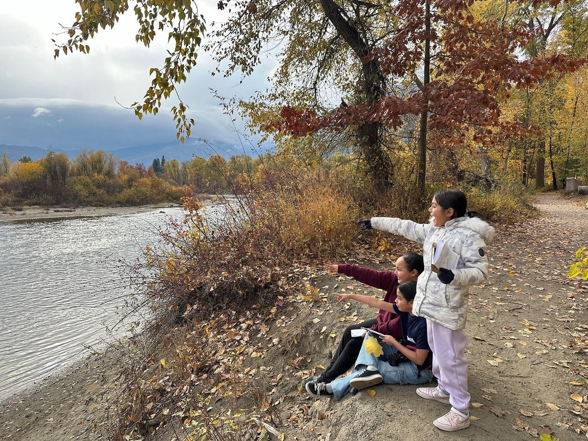A group of 3 girls sitting on the banks of the Wenatchee River with colorful leaves surrounding them, pointing at wildlife across the river.