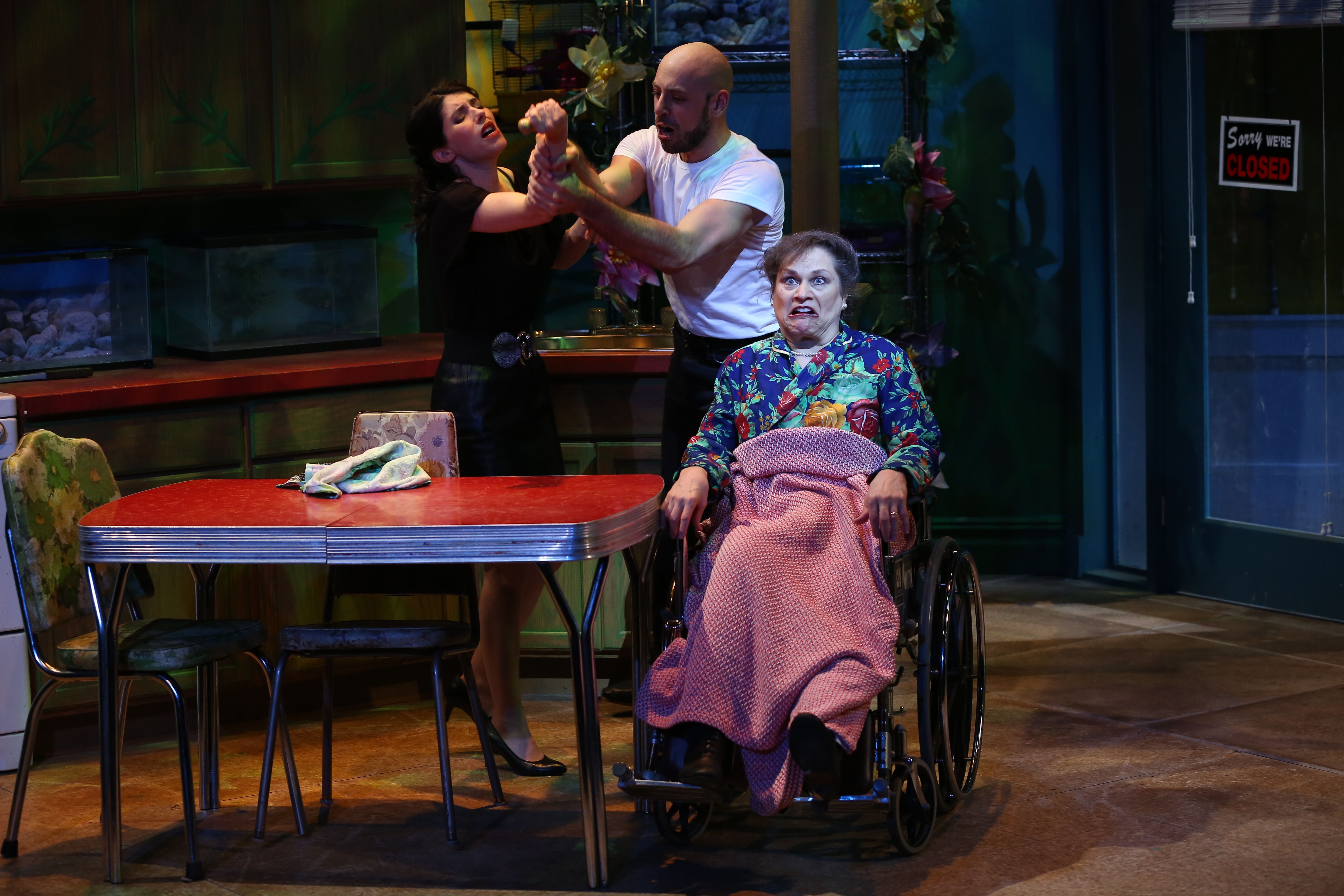 A picture of three actors who are performing another dark scene from The Artificial Jungle. One of the actors is sitting in a wheelchair and has a blanket on her lap. The other two actors are standing and arm wrestling over an unknown object. 