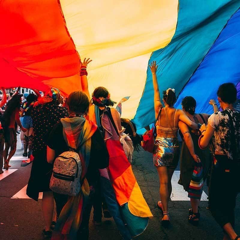 Many people with their hands up in the air walking below an oversized rainbow flag.