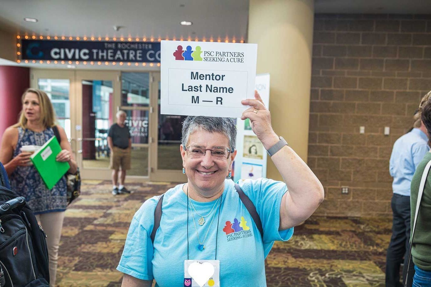 A grinning woman wearing a blue tshirt with a PSC Partners logo on it is holding a sign on her head. It reads Mentor Last Name M-R.