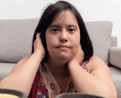 A woman with short dark hair is sitting on the floor in front of a beige sofa. She is leaning on a surface with her hands near her face. She is wearing a colorful patterned sleeveless top. Two out-of-focus cups or bowls are visible in the foreground.