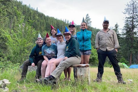 A crew sits on a bench posing for the camera, all making silly faces and wearing party hats.