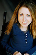 Shannon DeVido, a women in a power wheelchair. She is smiling widely and is wearing a blue blouse. The back drop is dark. She has long dark blonde hair. She is facing the camera directly.