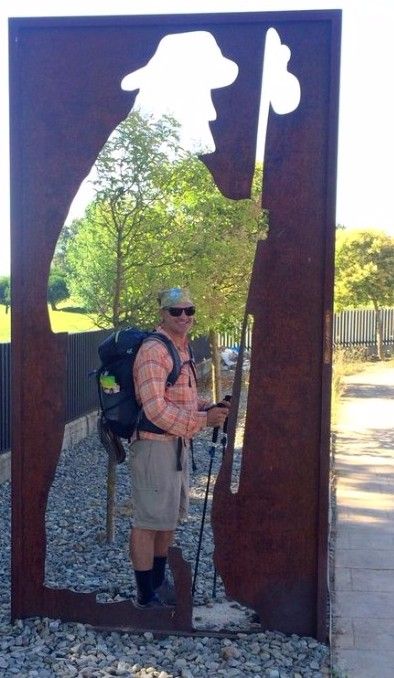 Man smiling, wearing a hiking backpack and caring hiking poles stands in a silhouette sculpture of a man with a hiking pole.