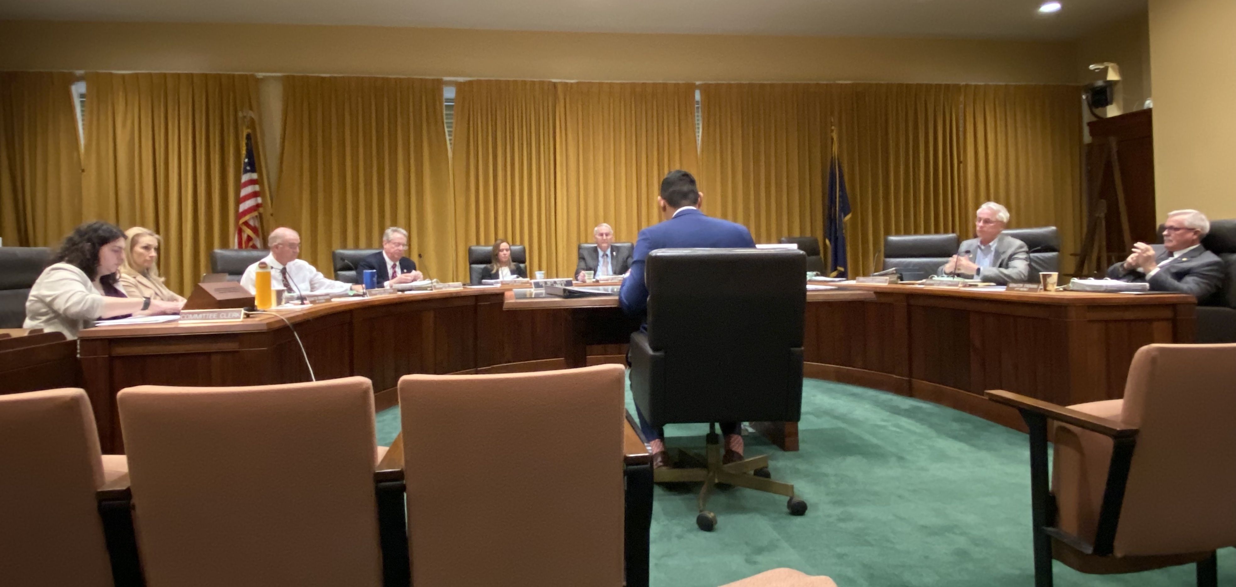 Senator Tony Vargas speaks before the Appropriations Committee of the Nebraska State Legislature. The back wall is floor to ceiling gold curtains and the carpet is green.