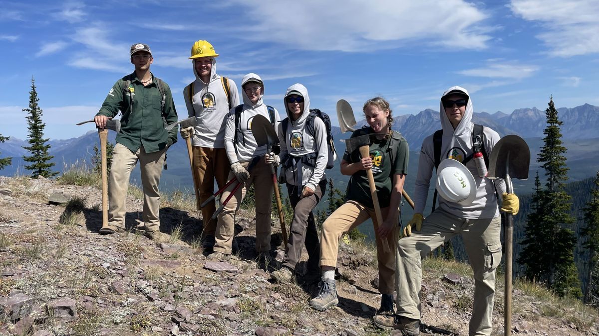 [Image Description: An MCC crew holds tools and poses on a ridgeline, mountains behind them.]