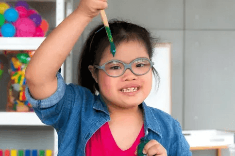 A young girl with glasses and a ponytail is holding a paintbrush covered in green paint above her head. She is wearing a denim shirt over a bright pink top. The background shows colorful toys and art supplies on shelves, indicating a creative setting.