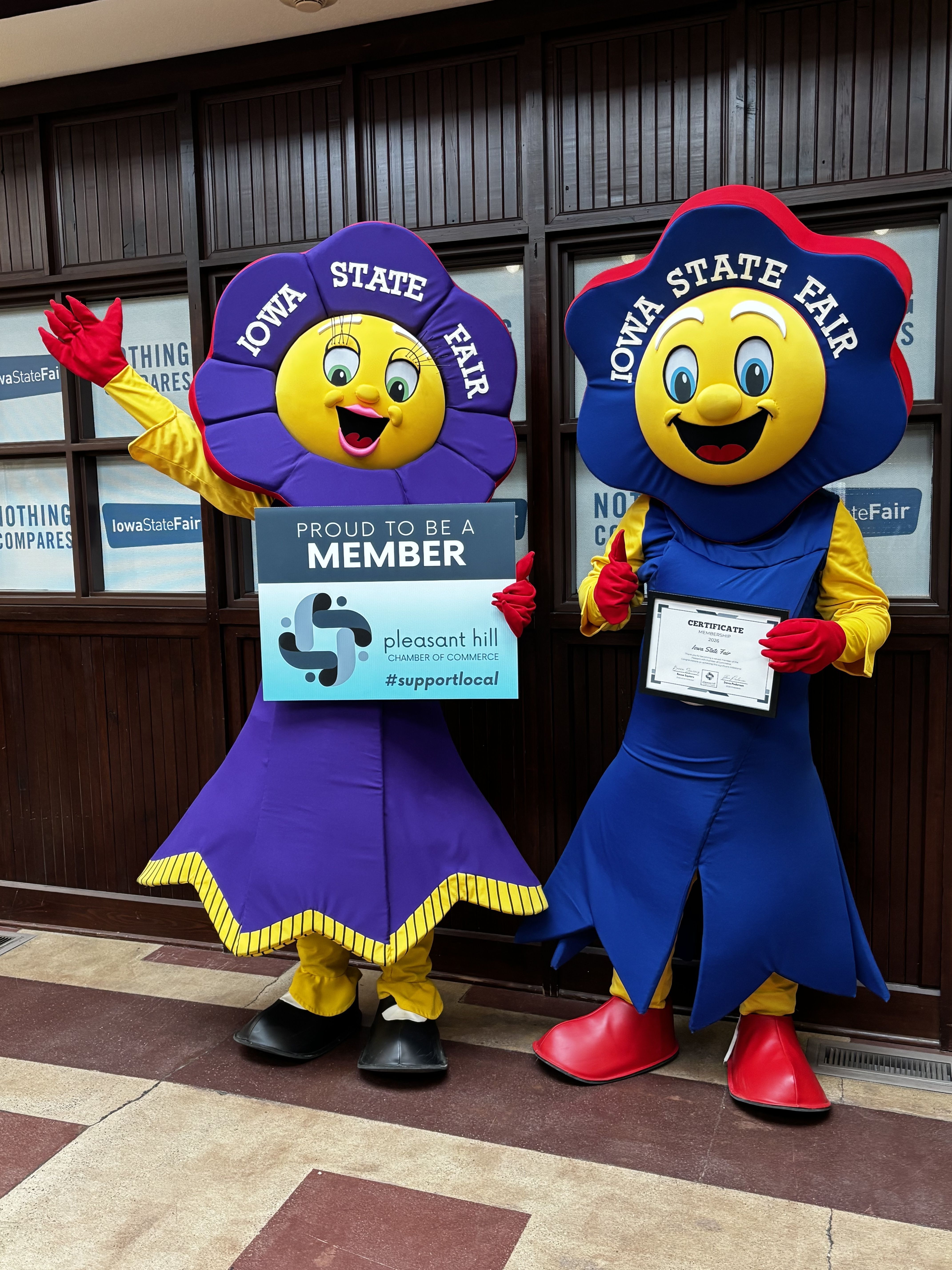 Two people in large, colorful award‑ribbon costumes stand outside an Iowa State Fair display, holding a certificate and a “Proud to be a member Pleasant Hill Chamber of Commerce” sign.