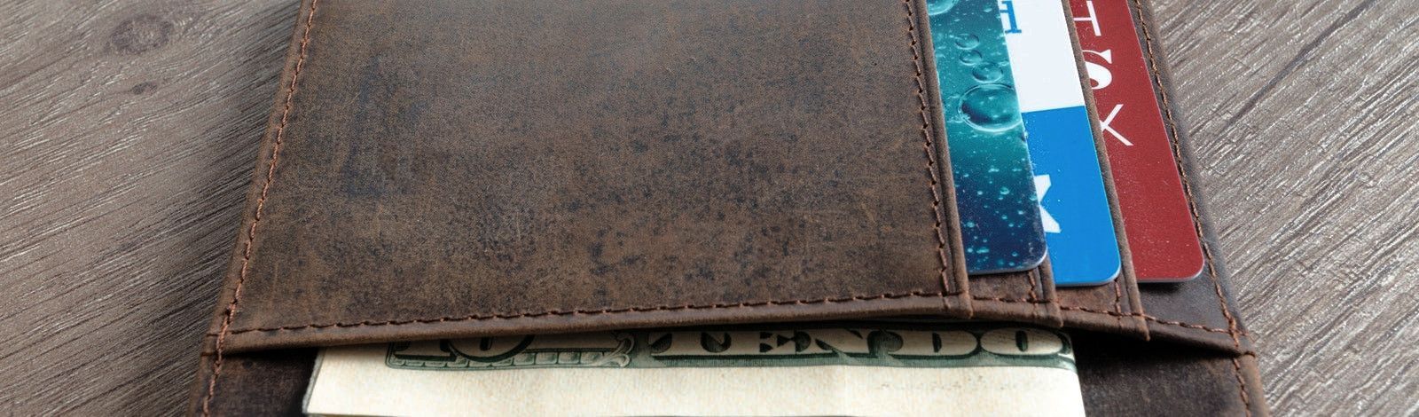 Closeup photo of a brown leather wallet with three credit cards and a ten dollar bill showing. The credit cards stick out of slots on the right side of the wallet; they are teal, blue and white, and red and white. The edge of the ten dollar bill sticks ou
