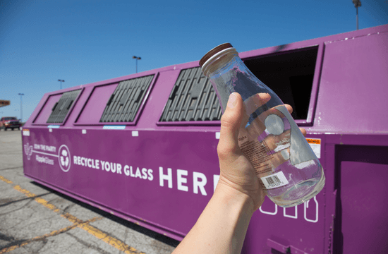 a glass bottle is held up in front of a purple glass-recycling container