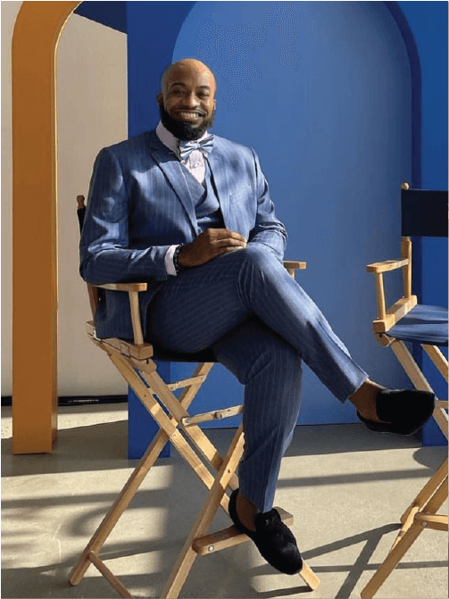 Steven McCoy is sitting in a chair with his legs crossed wearing a 3 piece blue suit