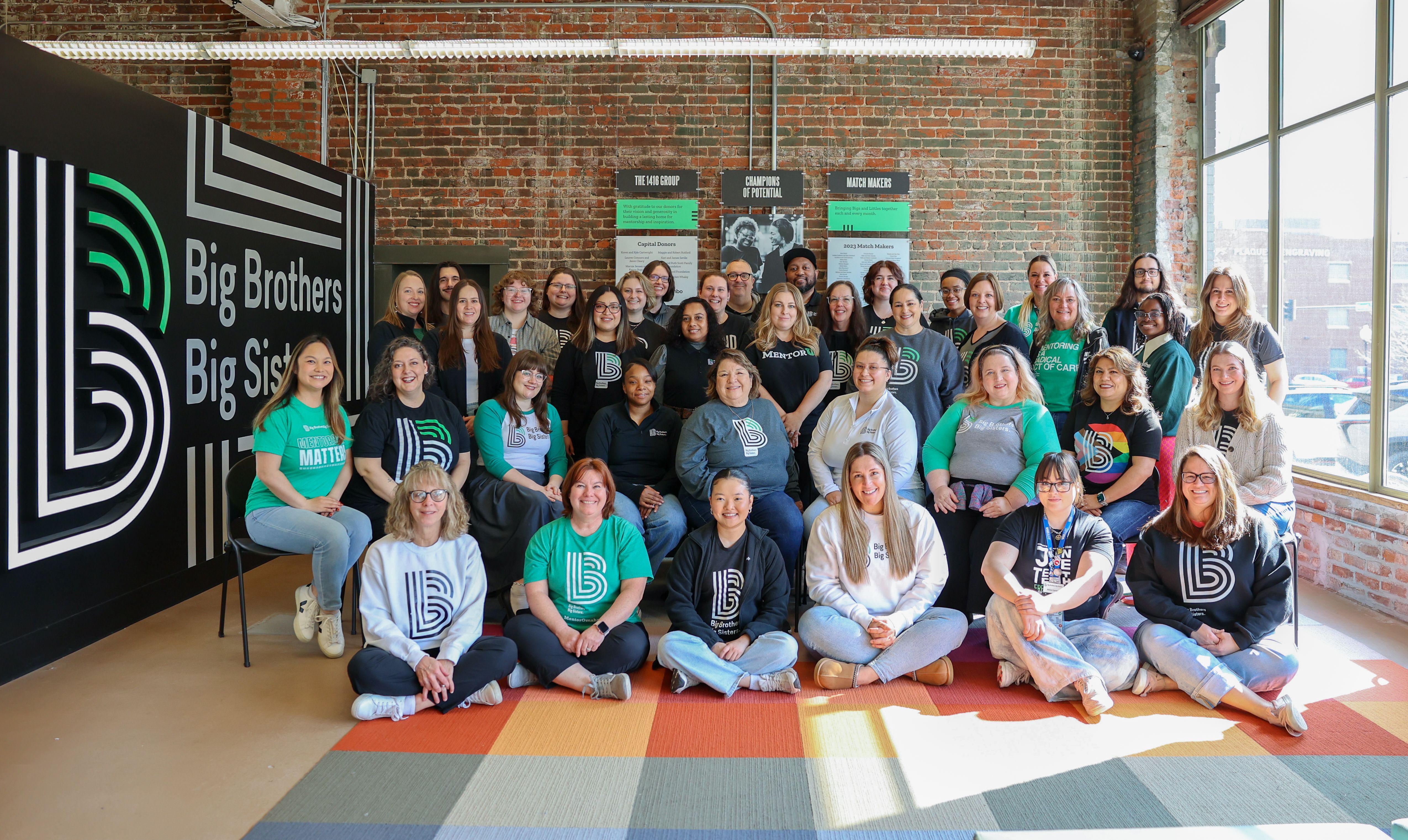 Group photo of Big Brothers Big Sisters of the Midlands staff gathered inside their office, smiling together in front of a large BBBS logo wall. Team members wear organization-branded shirts and sweatshirts in green, black, and gray, representing unity an