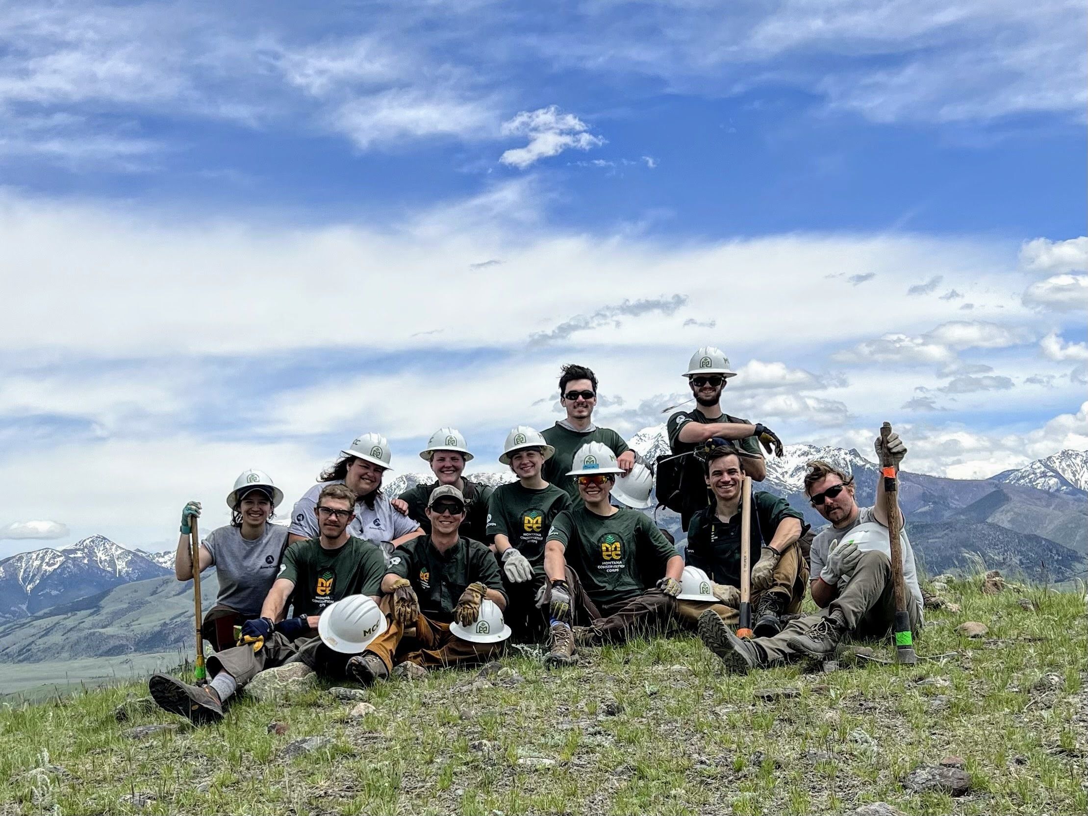 A group sits at an overlook, with white MCC leader helmets in their hands. There is a bright blue sky behind them.