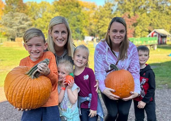 2 sisters stand with their 4 children, everyone holding pumpkins and grinning widely