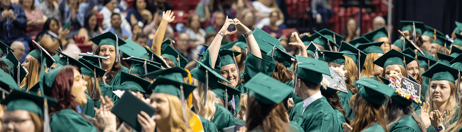 Many people dresses in graduation regalia celebrating in center is one student cupping hands together in heart shape above head and smiling.