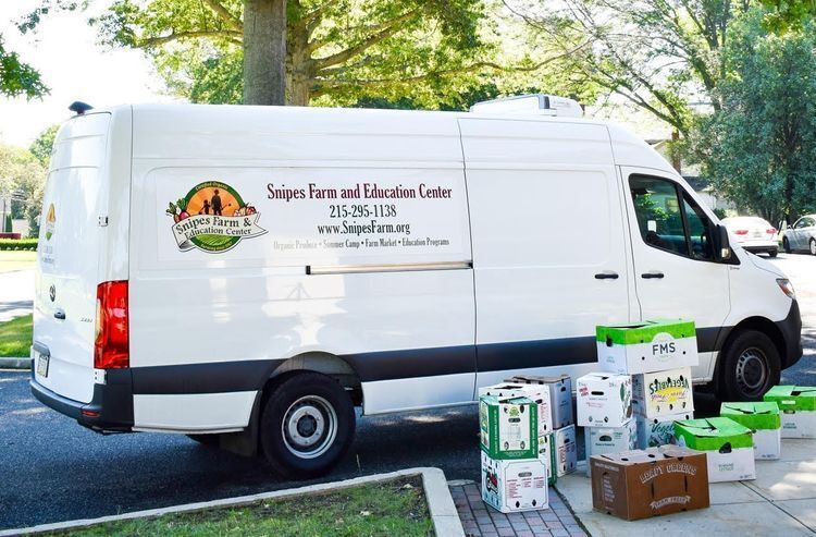 A big white van sits at the side of the curb with about 10 boxes of vegetables sitting in front of it.