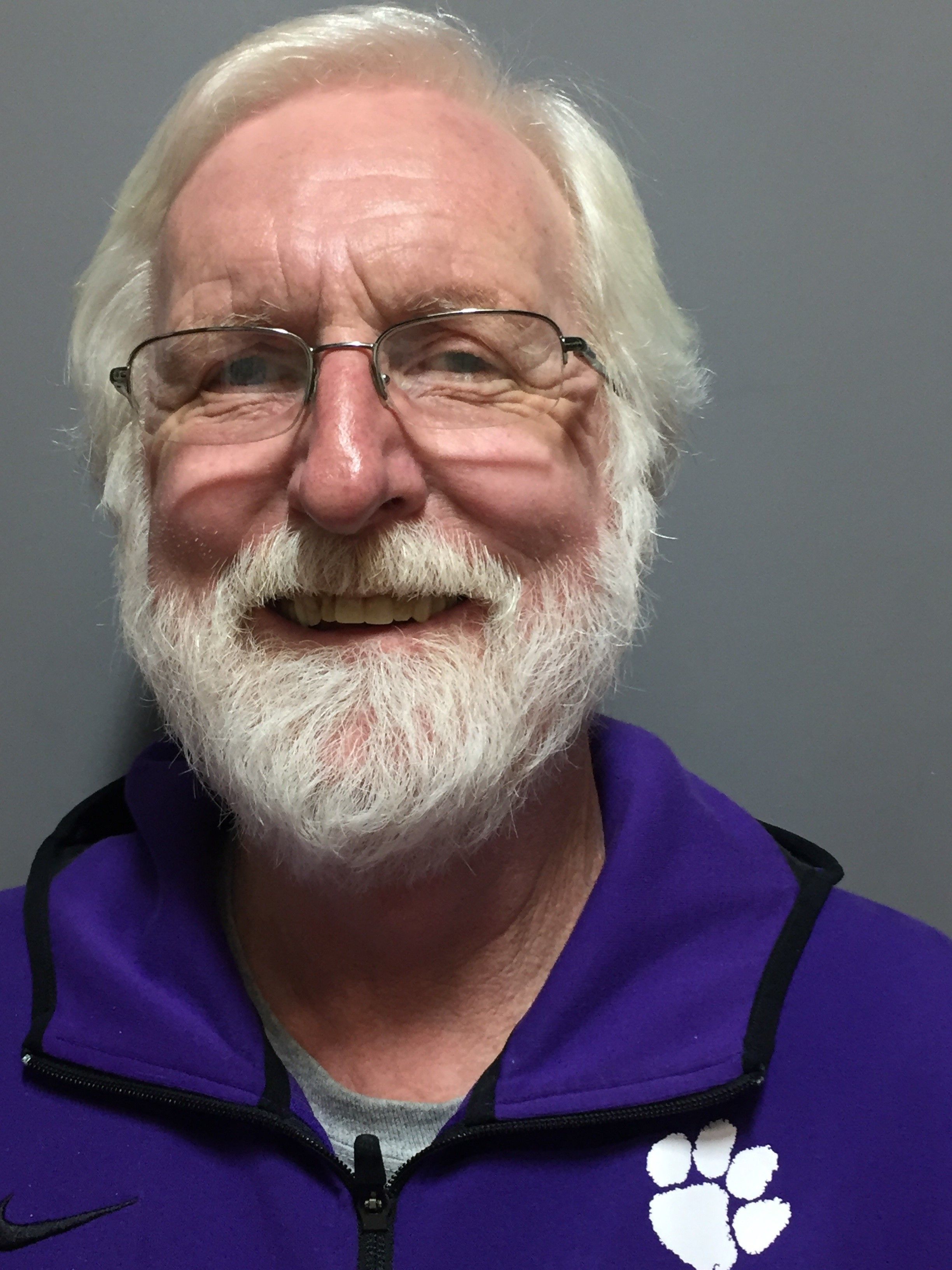 A white man with a white beard in a purple collared shirt and glasses smiles at the camera. This is Chris Jensen.