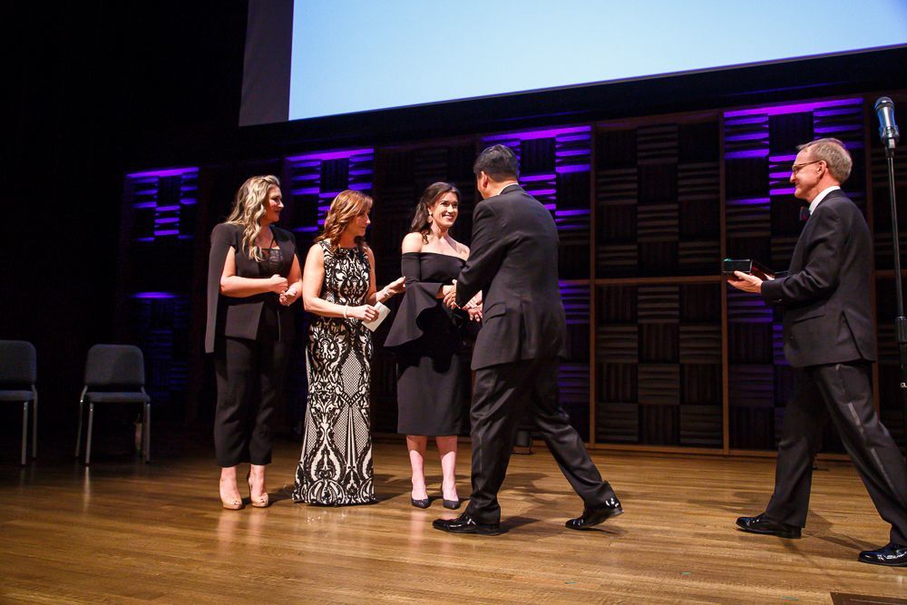 Rebecca Alexander, Tori Banu, and Nancy Corderman all stand on stage as Rebecca shakes hands with Suber and accepts her metal.