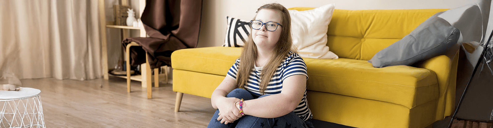 A young girl with Down syndrome sits on the floor in a bright room with a yellow couch behind her. She wears glasses, a striped shirt, and jeans. A small white side table and decorative elements like pillows and curtains are visible in the background.