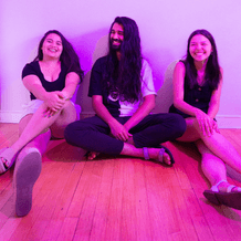 Three Asian American musicians sitting next to each with their backs again a plain white wall on a wood floor