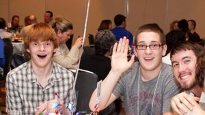 Three young adult males sit side-by-side smiling widely at the camera. One is waving. Conference goers can be seen sitting at other tables behind them.