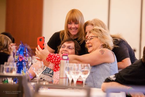 Four women stand huddled together taking a selfie. Around them are conference goers.