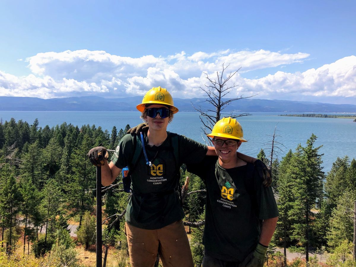 [Image Description: Two MCC youth members stand arm in arm, smiling, standing on a hillside that slopes down into a forest and glassy blue lake.]