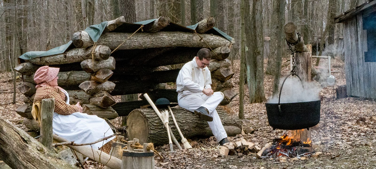 Two historical interpreters demonstrate boiling maple sap in an iron kettle near a log-crib lean-to.