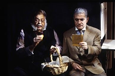 Jerry has a grey beard and is wearing a black vest with a plaid shirt, he's sitting on a chair in dim lighting, holding a basket while eating something. George is sitting next to Jerry in a lighter lighting while reading a paper. 