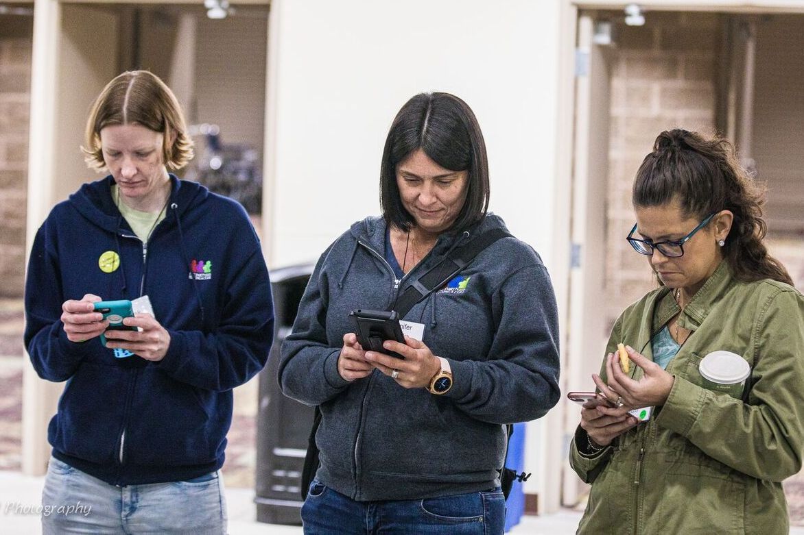 Three women are standing in a line looking at their phones. One is wearing a blue PSC Partners sweatshirt